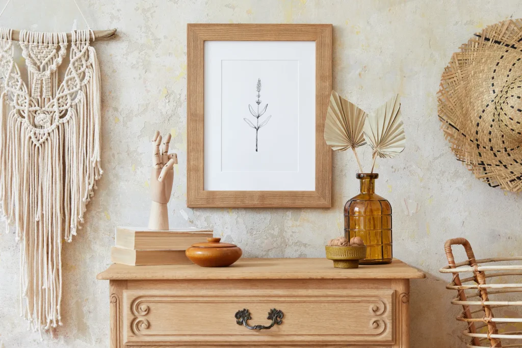 Holzkommode mit Dekoration im Boho-Stil vor strukturierter Wand: Makramee-Wandbehang, Holzhandmodell, Trockenpflanzen in Glasvase, Bücher, Holzdose und gerahmte botanische Zeichnung. Ideal für ein stilvoll eingerichtetes Tiny House.