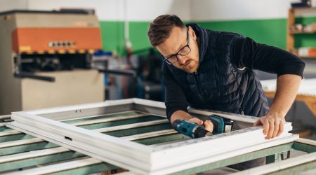 Handwerker montiert ein Fensterrahmenprofil in der Werkstatt und nutzt eine Bohrmaschine zur präzisen Befestigung.