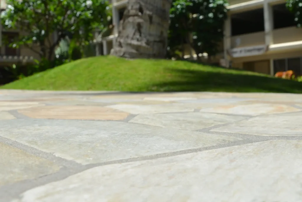 Nahaufnahme einer hellen, natursteinartigen Terrasse im Außenbereich mit polygonaler Plattenstruktur. Im Hintergrund unscharf sichtbar: ein kleiner, begrünt angelegter Hügel mit Palmenstamm und weiteren tropischen Pflanzen sowie ein Gebäude mit Balkonen.