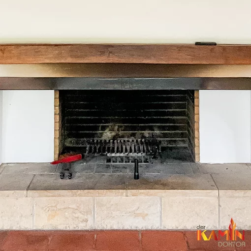 Offener gemauerter Kamin mit verrußter Brennkammer, breitem Holzrahmen und hellen Natursteinfliesen.
