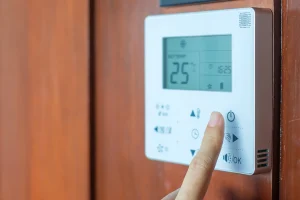 Finger drückt den Einschaltknopf an einem digitalen Raumthermostat mit Display zur Temperaturregelung an einer Holzwand.