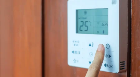 Finger drückt den Einschaltknopf an einem digitalen Raumthermostat mit Display zur Temperaturregelung an einer Holzwand.