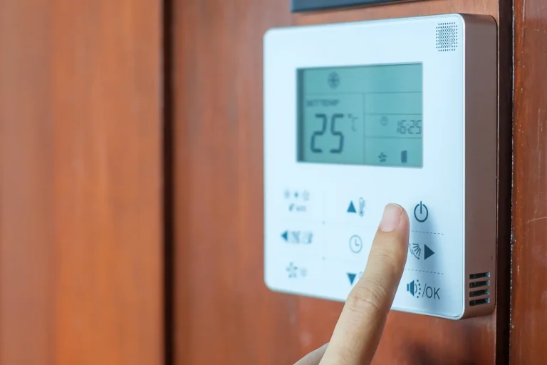Finger drückt den Einschaltknopf an einem digitalen Raumthermostat mit Display zur Temperaturregelung an einer Holzwand.