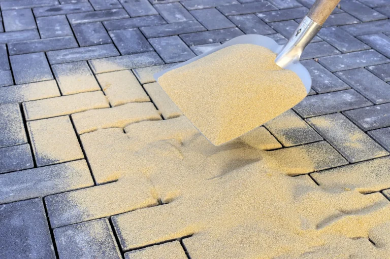 Ein Pflasterbelag aus dunkelgrauen Betonsteinen wird mit feinem, gelblichem Fugensand eingekehrt. Eine Metallschaufel verteilt den Sand auf der Fläche, um die Fugen zu stabilisieren.