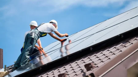 Zwei Solateure mit Schutzhelmen und Arbeitskleidung montieren Photovoltaikmodule auf einem geneigten Ziegeldach bei Tageslicht.