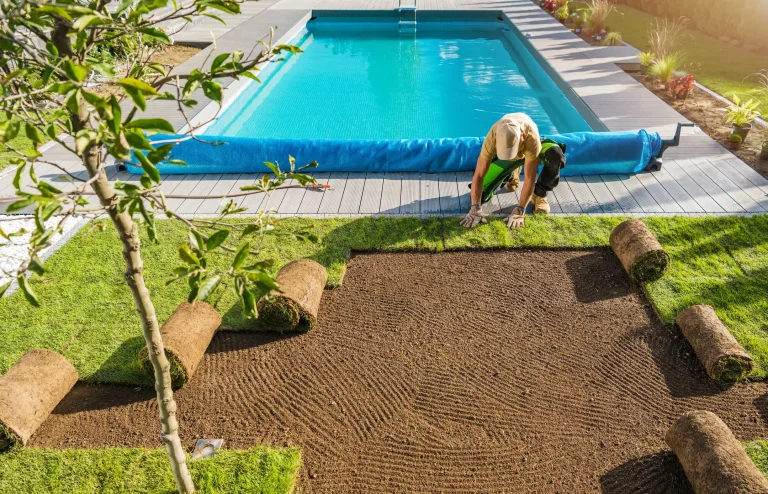 Ein Gärtner verlegt Rollrasen an einem frisch vorbereiteten Boden direkt neben einem Pool in einem modernen Garten. Mehrere aufgerollte Rasensegmente liegen bereit.