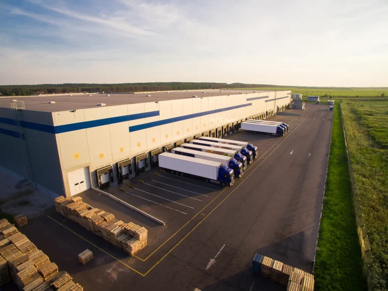 Großes Logistikzentrum mit mehreren Lkw an den Verladerampen und gestapelten Paletten auf dem Hof, umgeben von weiten Grünflächen.