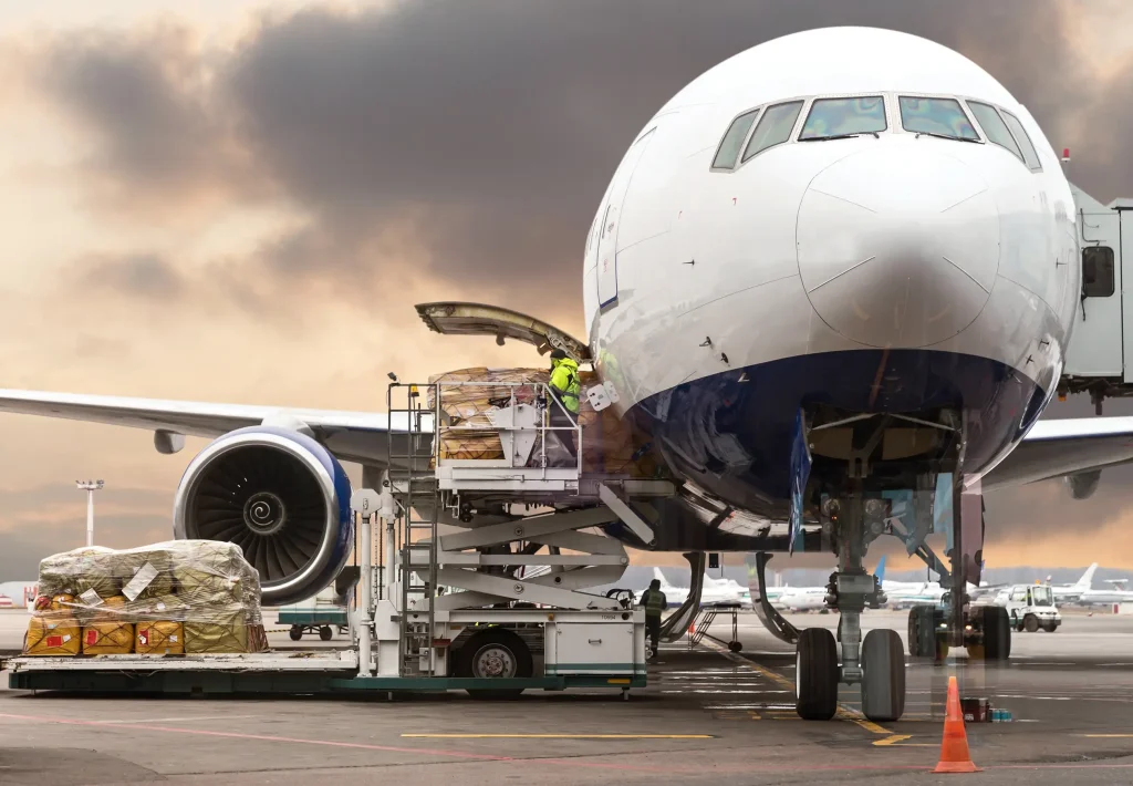 Frachtverladung an ein Verkehrsflugzeug am Flughafen bei Sonnenuntergang. Bodenpersonal lädt Paletten mit Waren in das vordere Frachtraumsegment eines großen Passagierflugzeugs.