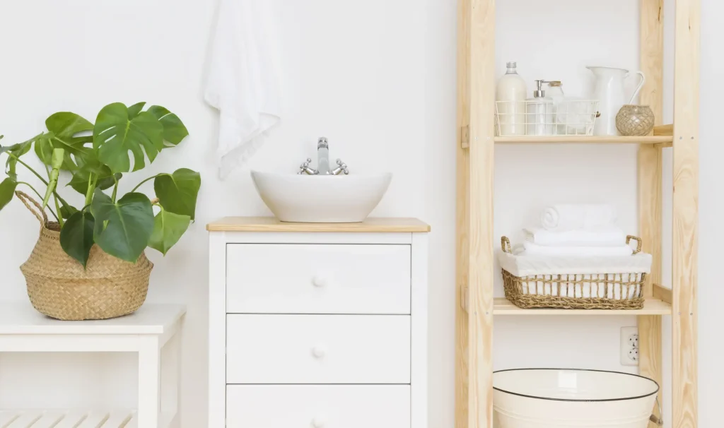 Waschplatz in einem Tiny House Bad mit rundem Aufsatzbecken auf weißem Unterschrank, daneben ein Regal mit Handtüchern und Pflegeprodukten sowie eine große Zimmerpflanze in einem Korb.