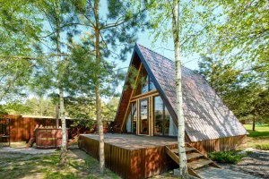 Tiny House in A-Frame-Bauweise mit großer Holzterrasse steht im Wald zwischen Birken und Kiefern. Links im Bild ist ein runder Hot Tub aus Holz zu sehen.
