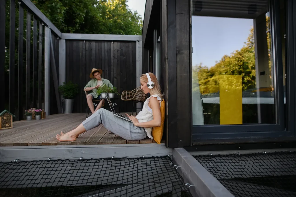 Junges Pärchen sitzt auf einer Holzterrasse vor einem dunklen Tiny House. Eine junge Frau sitzt mit einem Laptop und Kopfhörern an der Wand, während der junge Mann im Hintergrund mit Strohhut ein Buch liest.