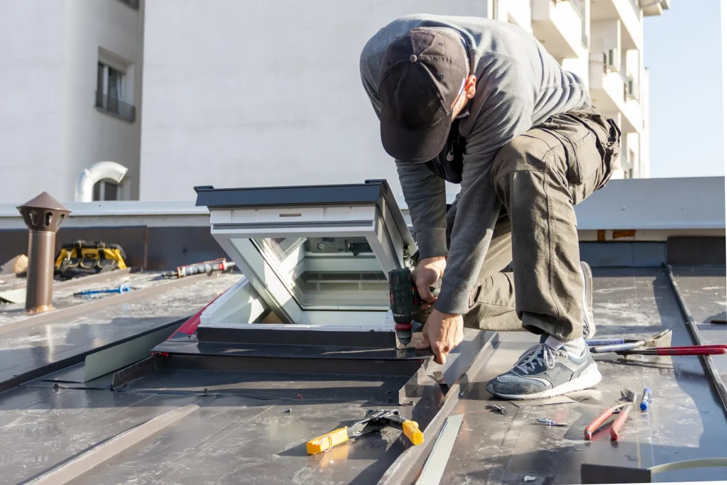 Ein Handwerker kniet auf einem Flachdach und dichtet ein neu eingebautes Dachfenster mit Werkzeugen und Abdichtungsmaterial ab. Im Hintergrund sind moderne Wohngebäude zu sehen.