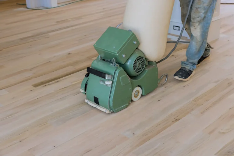 Handwerker schleift mit einer grünen Parkettschleifmaschine einen hellen Holzboden ab. Die Maschine hat einen beigefarbenen Staubfangsack, und im Hintergrund ist der unbehandelte Holzdielenboden sichtbar.