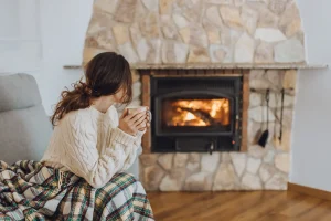 Frau mit lockigem Haar sitzt auf einem Sofa vor einem brennenden Kamin. Sie trägt einen cremefarbenen Strickpullover, ist in eine karierte Decke gehüllt und hält eine Tasse in beiden Händen. Der Kamin ist in eine Natursteinverkleidung eingefasst.