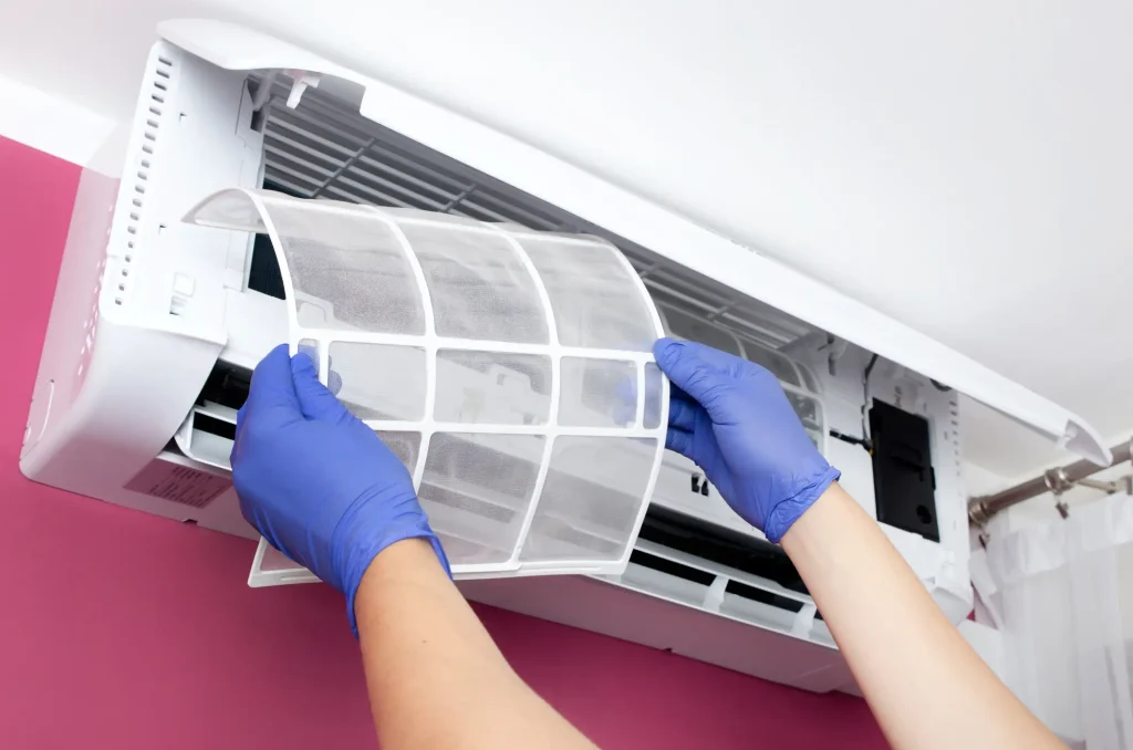 Ein Klimatechniker mit blauen Schutzhandschuhen entnimmt den Filter einer an der Wand montierten Klimaanlage zur Reinigung.