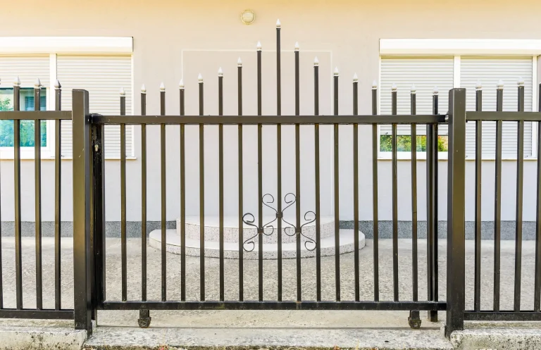Metallzaun mit senkrechten schwarzen Stäben und weißen Spitzen vor einem beige gestrichenen Gebäude mit zwei geschlossenen Rollladenfenstern und einem runden Treppenpodest in der Mitte.