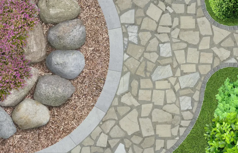 Gartenweg aus unregelmäßig verlegten Natursteinplatten mit sauber verfugten Fugen. Links angrenzend ein Beet mit Ziersteinen, Mulch und blühenden Pflanzen, rechts ein sauber geschnittener Rasen mit Buchsbaumhecke.