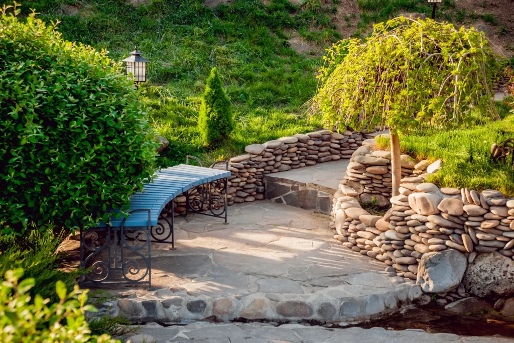 Gartenbereich mit gepflastertem Natursteinboden, geschwungenem Weg und gemörtelten Trockenmauern aus flachen Flusssteinen. Eine geschwungene blaue Gartenbank aus Metall steht zwischen üppigen Sträuchern und einem kleinen Baum. Die Fugen zwischen den Natursteinen sind sauber und harmonisch ausgeführt.