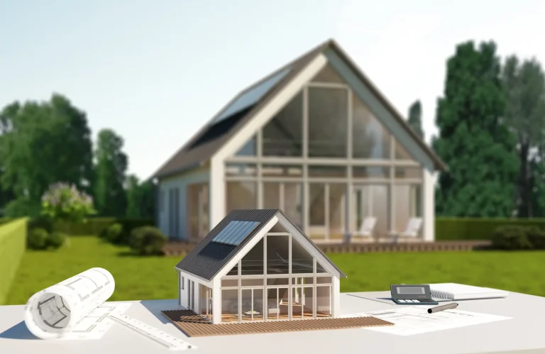 Architekturmodell eines modernen Einfamilienhauses mit Satteldach, großen Fensterfronten und Solarpanelen, im Vordergrund auf einem Planungstisch mit Bauplänen, Lineal, Stift und Taschenrechner, im Hintergrund das realisierte Gebäude in grüner Gartenumgebung.