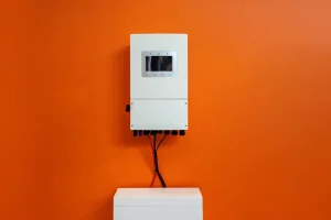 Weißer Wechselrichter mit Display oberhalb eines Batteriespeichersystems an einer orange gestrichenen Wand.