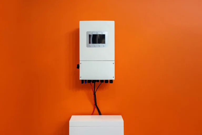Weißer Wechselrichter mit Display oberhalb eines Batteriespeichersystems an einer orange gestrichenen Wand.