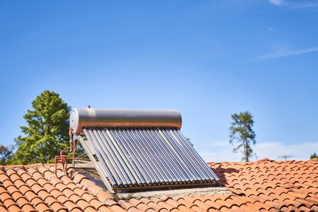 Solarkollektor mit Vakuumröhren auf einem roten Ziegeldach unter blauem Himmel, zur solaren Warmwasserbereitung installiert, mit Bäumen im Hintergrund.