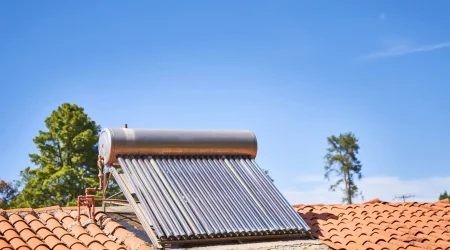 Solarkollektor mit Vakuumröhren auf einem roten Ziegeldach unter blauem Himmel, zur solaren Warmwasserbereitung installiert, mit Bäumen im Hintergrund.