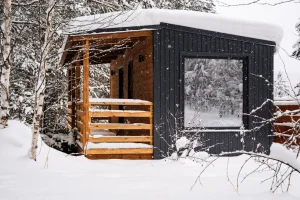 Kleines modernes Tiny House aus Holz mit schwarzer vertikaler Außenverkleidung und Flachdach, das von einer dicken Schneeschicht bedeckt ist. Das Gebäude steht in einer verschneiten Waldlandschaft. Die vordere Terrasse ist mit naturbelassenem Holz verkleidet, und ein großes Fenster spiegelt die winterliche Umgebung. Schneefall ist sichtbar, Bäume und Sträucher sind mit Schnee bedeckt.