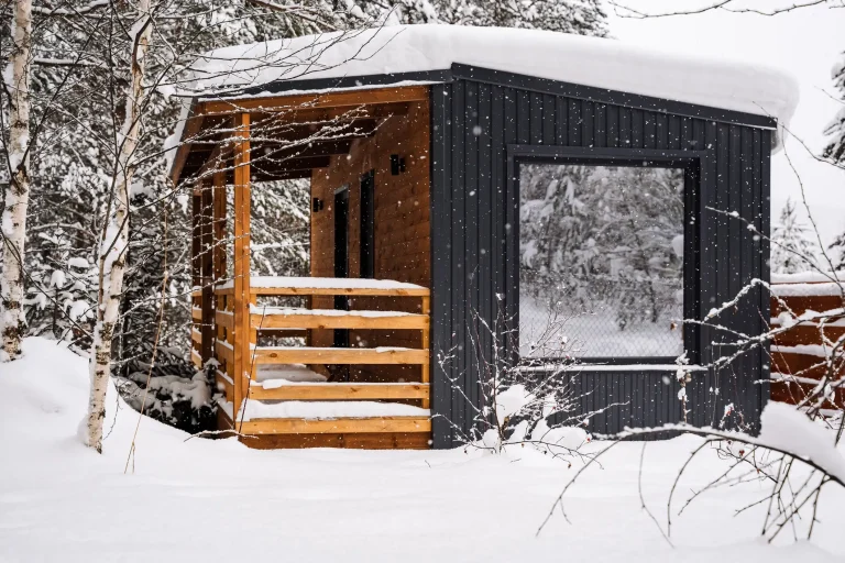 Kleines modernes Tiny House aus Holz mit schwarzer vertikaler Außenverkleidung und Flachdach, das von einer dicken Schneeschicht bedeckt ist. Das Gebäude steht in einer verschneiten Waldlandschaft. Die vordere Terrasse ist mit naturbelassenem Holz verkleidet, und ein großes Fenster spiegelt die winterliche Umgebung. Schneefall ist sichtbar, Bäume und Sträucher sind mit Schnee bedeckt.