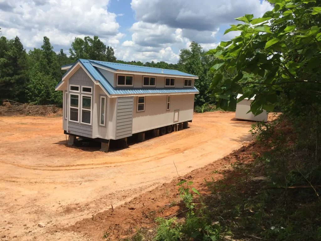 Ein Tiny House mit Satteldach und mehreren Fenstern steht auf einem Fahrgestell mit Rädern auf einer Baustelle, bereit zum Transport über die Straße.