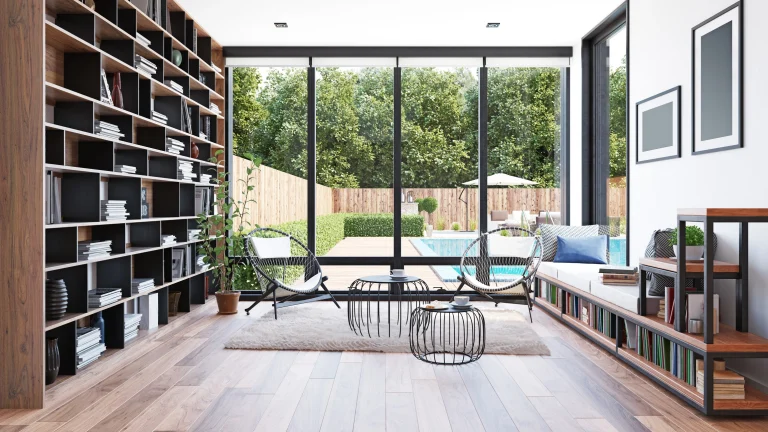Großes helles Wohnzimmer mit einer breiten Fensterfront über die ganze Wand, mit Blick in den Garten. Auf der linken Seite befindet sich ein Bücherregals aus braunem massiven Holz.