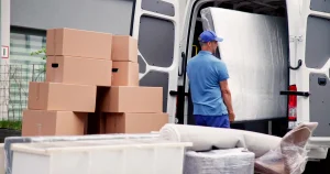 Ein Umzugshelfer lädt eine große Matratze in einen weißen Transporter, der mit offenen Türen am Straßenrand steht. Im Vordergrund liegen Teppiche, Möbel und gestapelte Umzugskartons bereit.