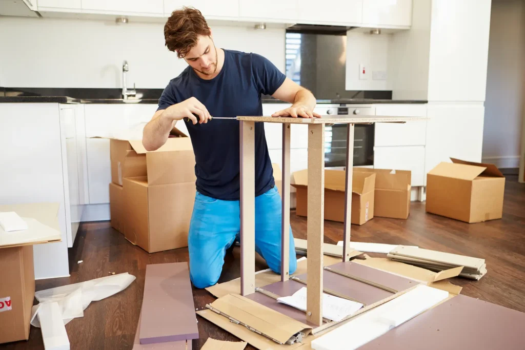 Ein Mann kniet in einer modernen Küche und montiert eigenständig ein Möbelstück aus Holzplatten. Um ihn herum stehen geöffnete Umzugskartons und Verpackungsmaterial.