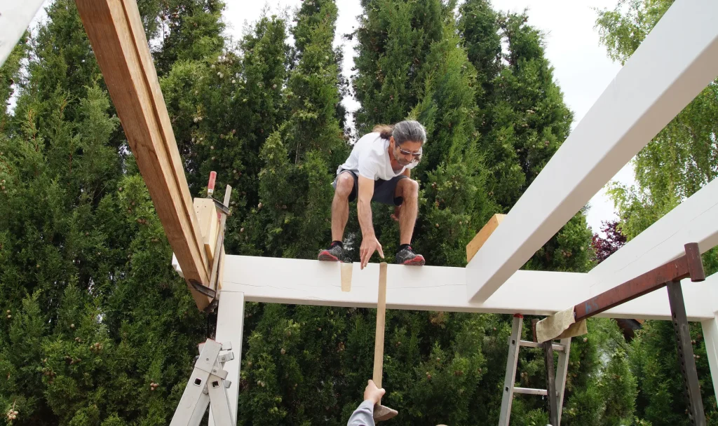 Ein Mann sitzt auf einem weißen Holzbalken einer Carport-Konstruktion in luftiger Höhe und greift nach einem Hammer, der ihm von unten gereicht wird. Im Hintergrund dichte, hohe Tannen.