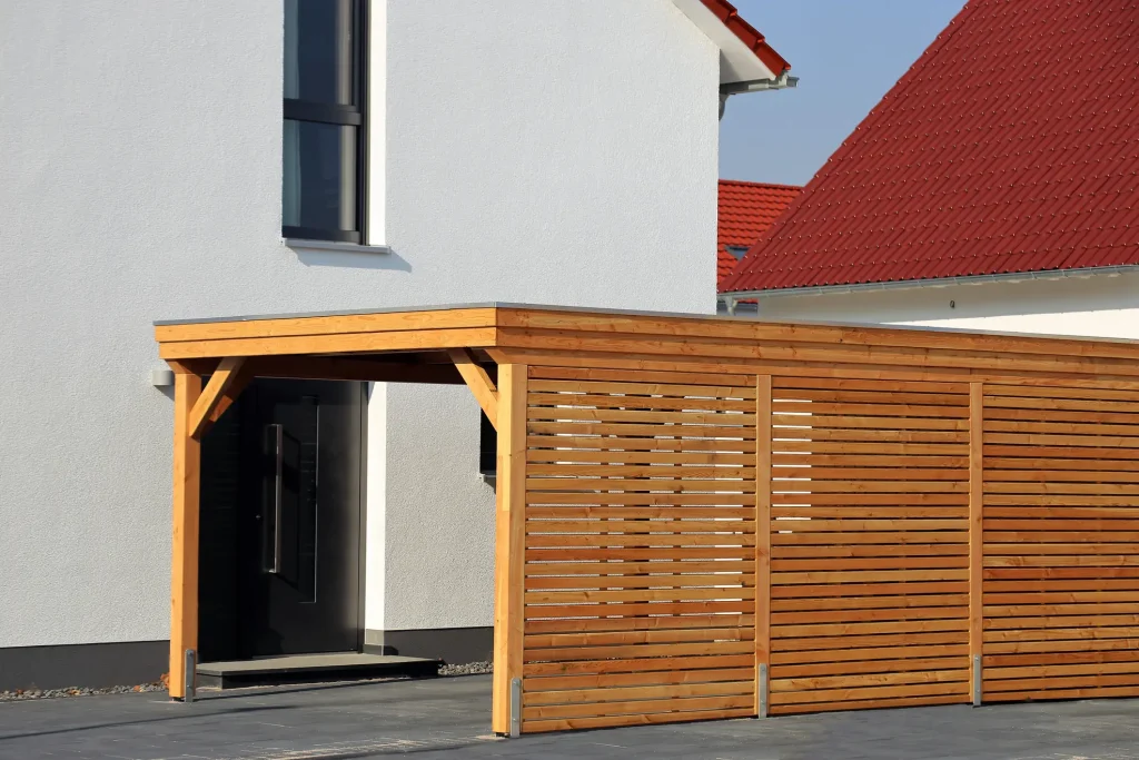 Carport mit Flachdach aus Holz, inklusive seitlicher Sichtschutzwand aus horizontalen Holzlamellen.