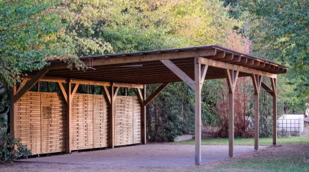 Holz Carport mit Flachdach und verkleideter, seitlicher Sichtschutzwand, auf Pflasterfläche im Grünen errichtet.