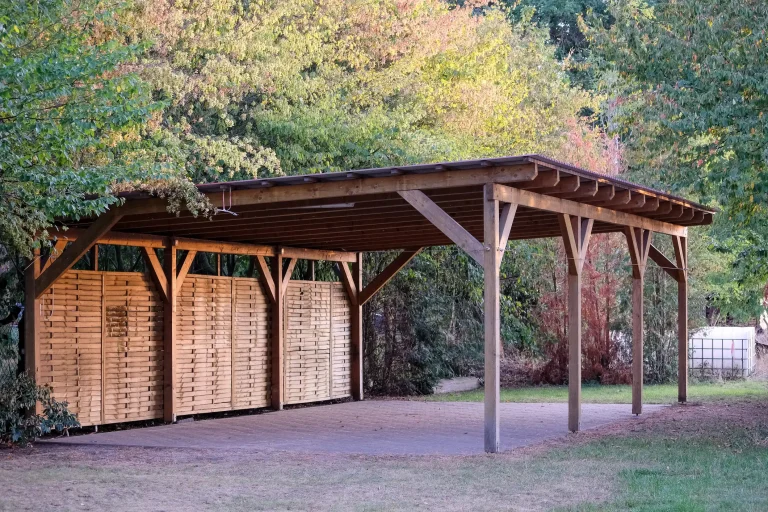 Holz Carport mit Flachdach und verkleideter, seitlicher Sichtschutzwand, auf Pflasterfläche im Grünen errichtet.