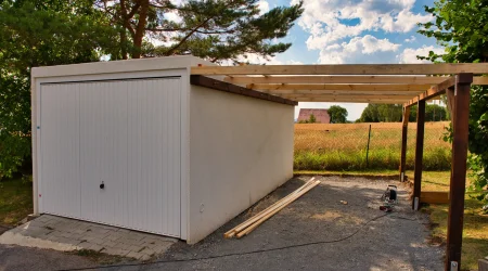 Teilweise überdachter Carport-Anbau aus Holz neben einer weißen Fertiggarage in ländlicher Umgebung. Die Konstruktion ist noch im Bau, Werkzeuge und Baumaterial liegen auf dem Boden. Im Hintergrund Felder, Bäume und ein Hausdach unter bewölktem Himmel.