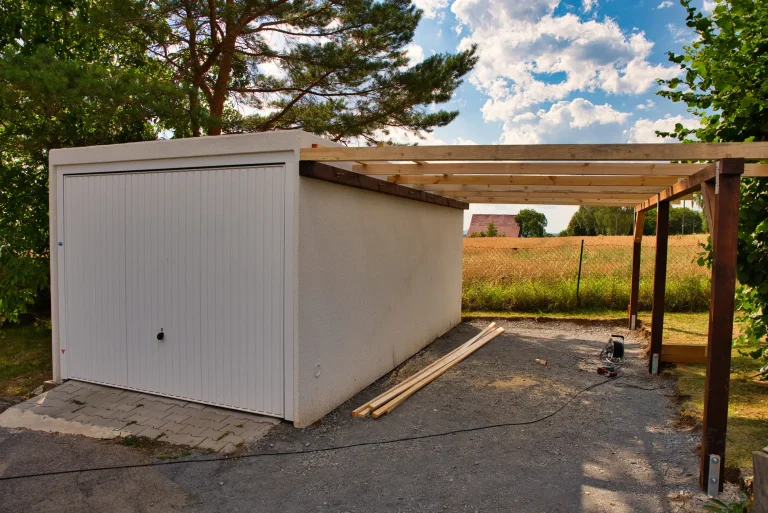 Teilweise überdachter Carport-Anbau aus Holz neben einer weißen Fertiggarage in ländlicher Umgebung. Die Konstruktion ist noch im Bau, Werkzeuge und Baumaterial liegen auf dem Boden. Im Hintergrund Felder, Bäume und ein Hausdach unter bewölktem Himmel.