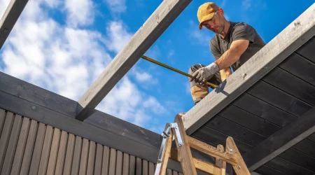 Handwerker auf einer Leiter misst mit einem Maßband Holzbalken eines Carportdaches ab, blauer Himmel mit Wolken im Hintergrund.