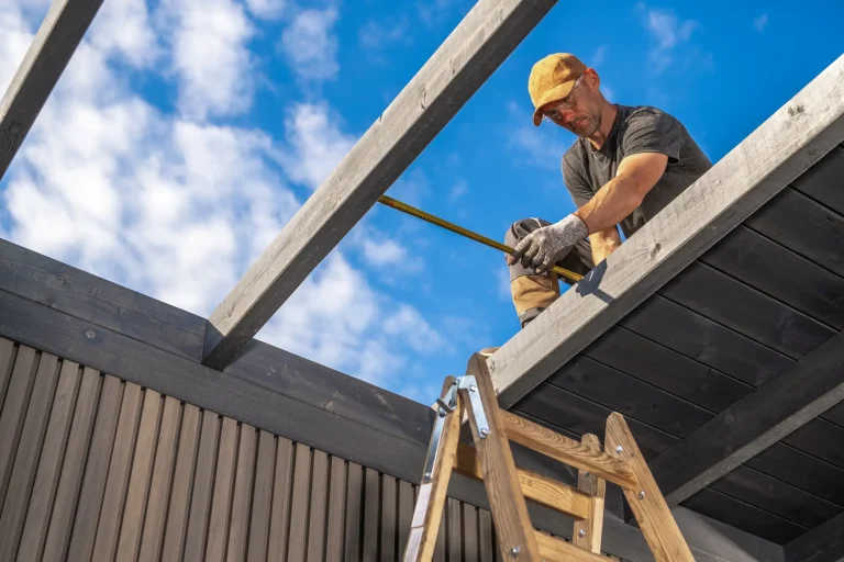 Handwerker auf einer Leiter misst mit einem Maßband Holzbalken eines Carportdaches ab, blauer Himmel mit Wolken im Hintergrund.