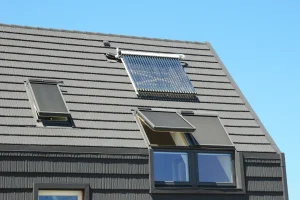 Modernes Satteldach mit dunkelgrauen Ziegeln, ausgestattet mit zwei Kniestockfenstern, die außenliegende Sonnenschutzrollos tragen. Oberhalb der Fenster ist eine solarthermische Anlage mit Vakuumröhrenkollektoren montiert. Im Hintergrund ist ein klarer blauer Himmel zu sehen.