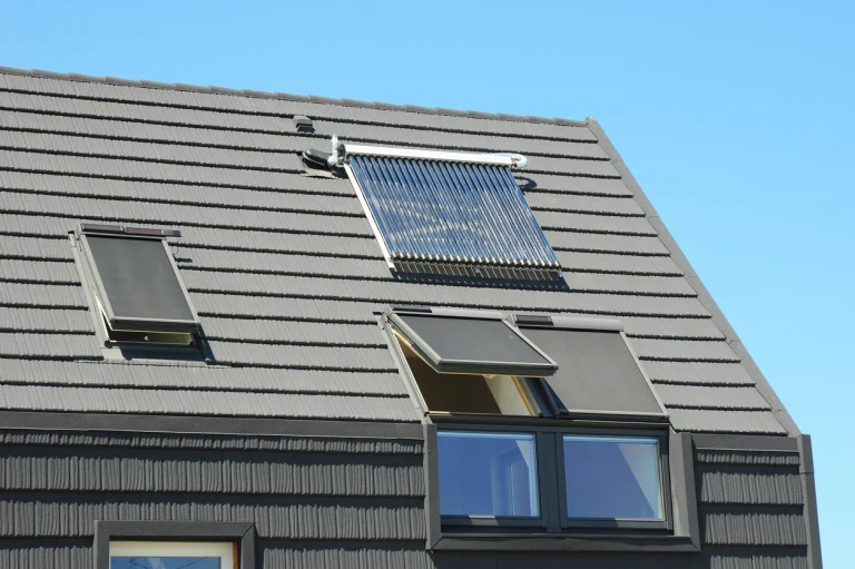 Modernes Satteldach mit dunkelgrauen Ziegeln, ausgestattet mit zwei Kniestockfenstern, die außenliegende Sonnenschutzrollos tragen. Oberhalb der Fenster ist eine solarthermische Anlage mit Vakuumröhrenkollektoren montiert. Im Hintergrund ist ein klarer blauer Himmel zu sehen.