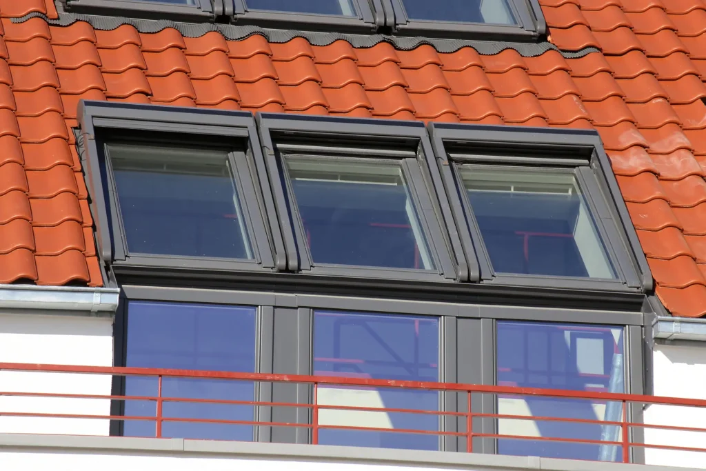 Drei nebeneinander liegende Kniestockfenster in einem schrägen Dach mit roten Ziegeln. Die Fenster sind modern gerahmt und gehören zu einem Dachgeschoss mit vorgelagerter Glasfassade und rotem Geländer.