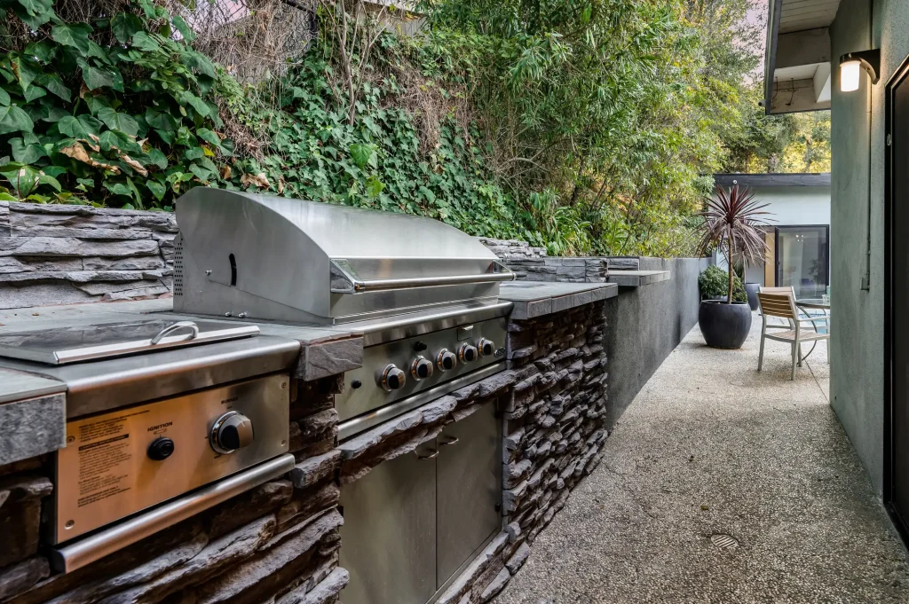 Eingelassene Outdoor-Küche mit Edelstahl-Gasgrill auf einer Terrasse. Die Küchenzeile ist mit dunklem Naturstein verkleidet. Im Hintergrund: Sitzbereich und grüne Bepflanzung.