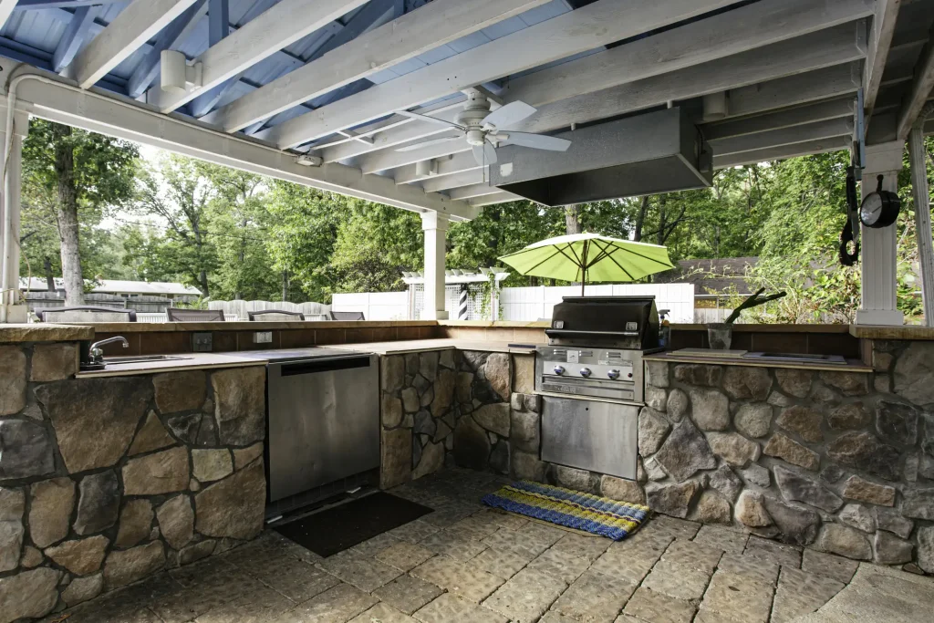 Überdachte Outdoor-Küche mit gemauerter Theke aus Naturstein, eingebautem Edelstahlgrill, Spüle und Deckenventilator. Im Hintergrund: Gartenbereich mit Sitzplätzen und grünem Sonnenschirm.