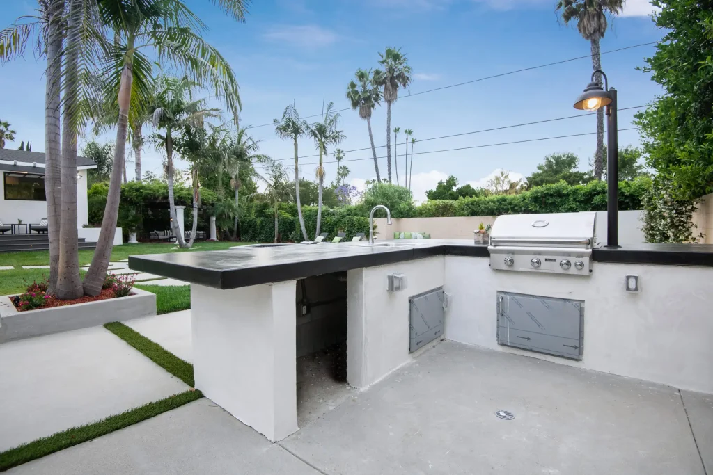 Moderne Outdoor-Küche mit weiß verputzter Theke, dunkler Arbeitsplatte, eingebautem Edelstahlgrill und Spüle, umgeben von Palmen und gepflegtem Garten.