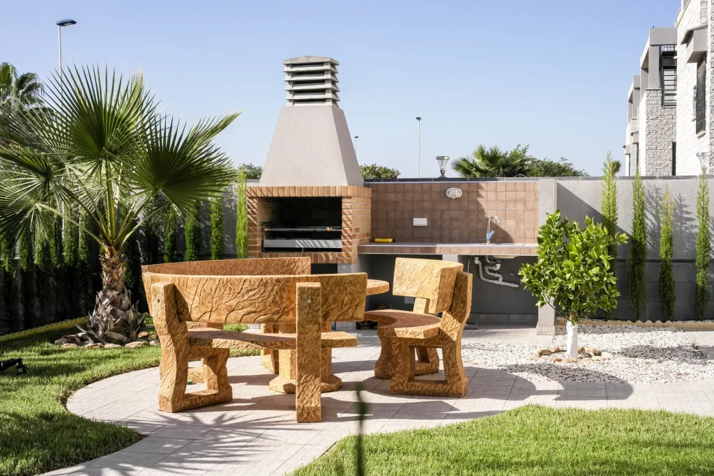 Gepflegter Garten mit gemauerter Grillstation, runder Sitzgruppe aus Holzimitat und angrenzender Außenküche mit Spüle und gefliestem Spritzschutz.