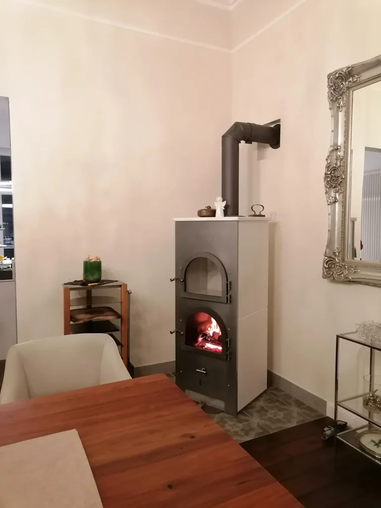Ein Römerofen Compact steht in einer modern eingerichteten Wohnung. Im unteren Fach brennt ein Feuer. Das Anschlussrohr führt seitlich in die Wand.