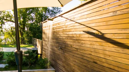 Warmwasser Außendusche mit senkrecht montierter Holzverkleidung, Wasser läuft aus einer schmalen Duscharmatur entlang der Holzwand, im Vordergrund Schirm, im Hintergrund Garten mit Grill.